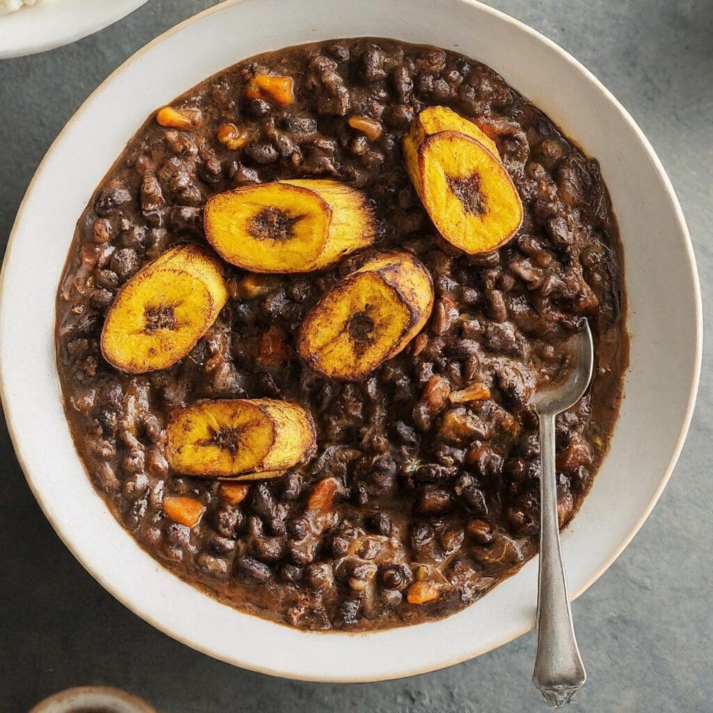 A steaming pot of Nigerian black bean stew with sliced plantains. The stew is garnished with chopped fresh herbs